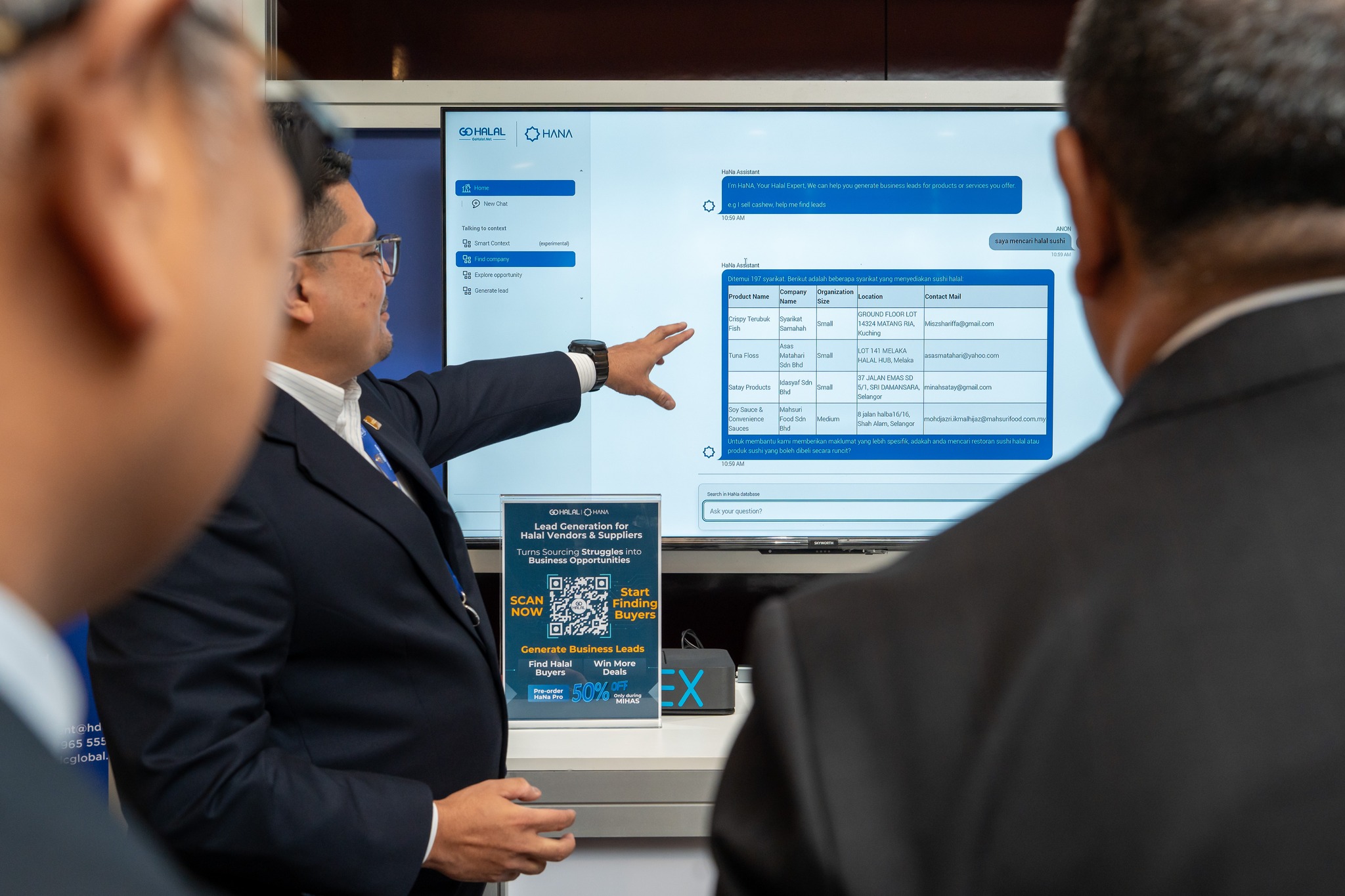 A professional demonstration of the HaNa AI sourcing interface on a large flat-screen display, with a business representative pointing out verified Halal supplier listings and contact data to corporate clients.